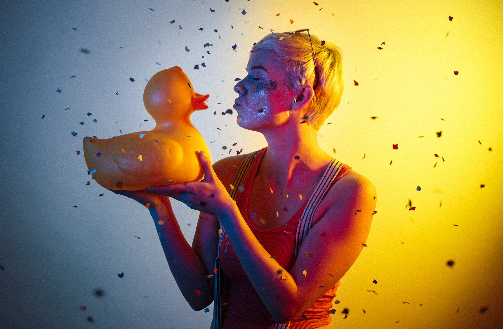 portrait of young woman with short hair wearing sunglasses and holding rubber duck with yellow and blue studio light